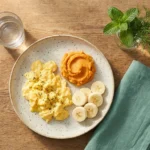 Small gastroparesis-friendly meal of scrambled eggs, mashed sweet potato, ripe banana, and water on a wooden table
