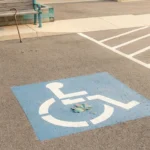 A clean accessible parking space with the international access symbol painted on blue asphalt, a cane resting against a nearby bench, and an autumn leaf — anchoring a respectful look at handicap parking permits for neuropathy.