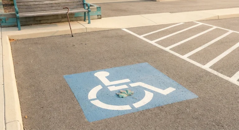 A clean accessible parking space with the international access symbol painted on blue asphalt, a cane resting against a nearby bench, and an autumn leaf — anchoring a respectful look at handicap parking permits for neuropathy.