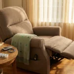 A taupe power lift recliner in a softly lit living room with sage throw, side table holding water, book, TENS unit, and pill organizer — anchoring a thoughtful chair choice for neuropathy.