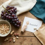 Overhead flat-lay of dark grapes, raw peanuts, a small vial of resveratrol powder, and supplement capsules on a wooden table