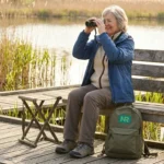 Woman in her sixties seated on a bench at a marsh boardwalk with binoculars raised, watching birds in golden morning light.