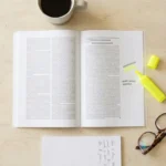 A printed journal article, highlighter, reading glasses and notepad arranged on a bright desk