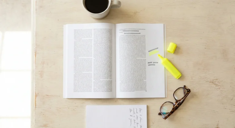 A printed journal article, highlighter, reading glasses and notepad arranged on a bright desk