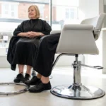 Woman seated in a salon chair with her feet flat on the floor beside the unused chrome footring.