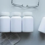 Four unlabelled pill bottles in a row on a grey desk, one pushed aside with its cap off