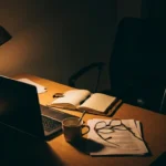 An empty desk chair and lamplit desk with notes and reading glasses at night